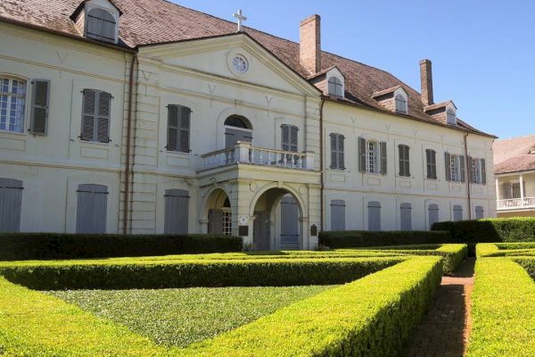 A large, classic villa with a symmetrical façade and arched entrance is surrounded by neatly trimmed hedges under a clear blue sky.